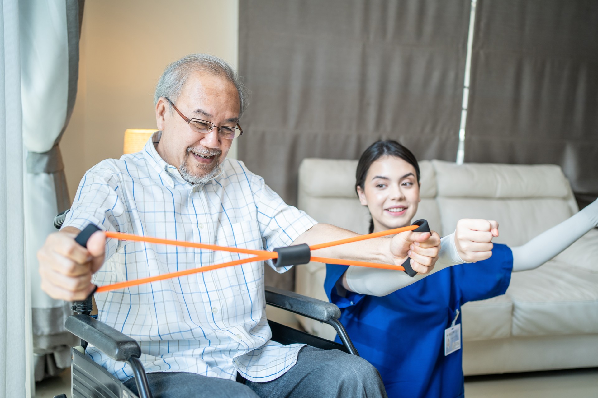 Asian Disabled senior elderly man on wheelchair doing physiotherapist with support from therapist nurse. Older handicapped man using resistance stretch band exercise for patient in home nursing care